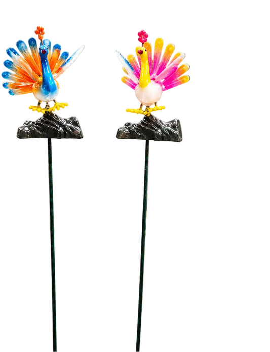 Two colorful garden stakes featuring a blue and orange peacock and a pink, yellow, and white peacock, each standing on a black rock base with a butterfly decoration to the left, set against a plain white background.