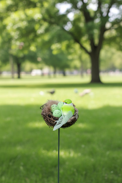 A decorative garden stake topped with a twig nest holding three speckled light green eggs and a bright yellow-green bird figurine with pale green wings, shown in front of a blurred wooden fence and potted plants.