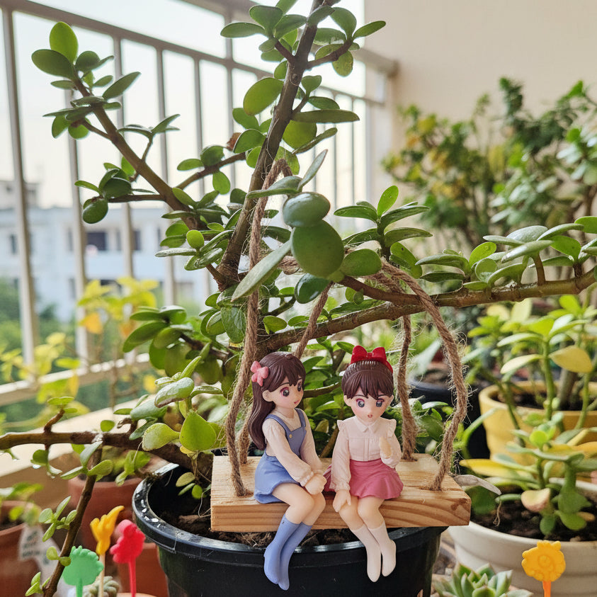 Two figurines of girls on a swing inside a potted plant with a blurred indoor background.